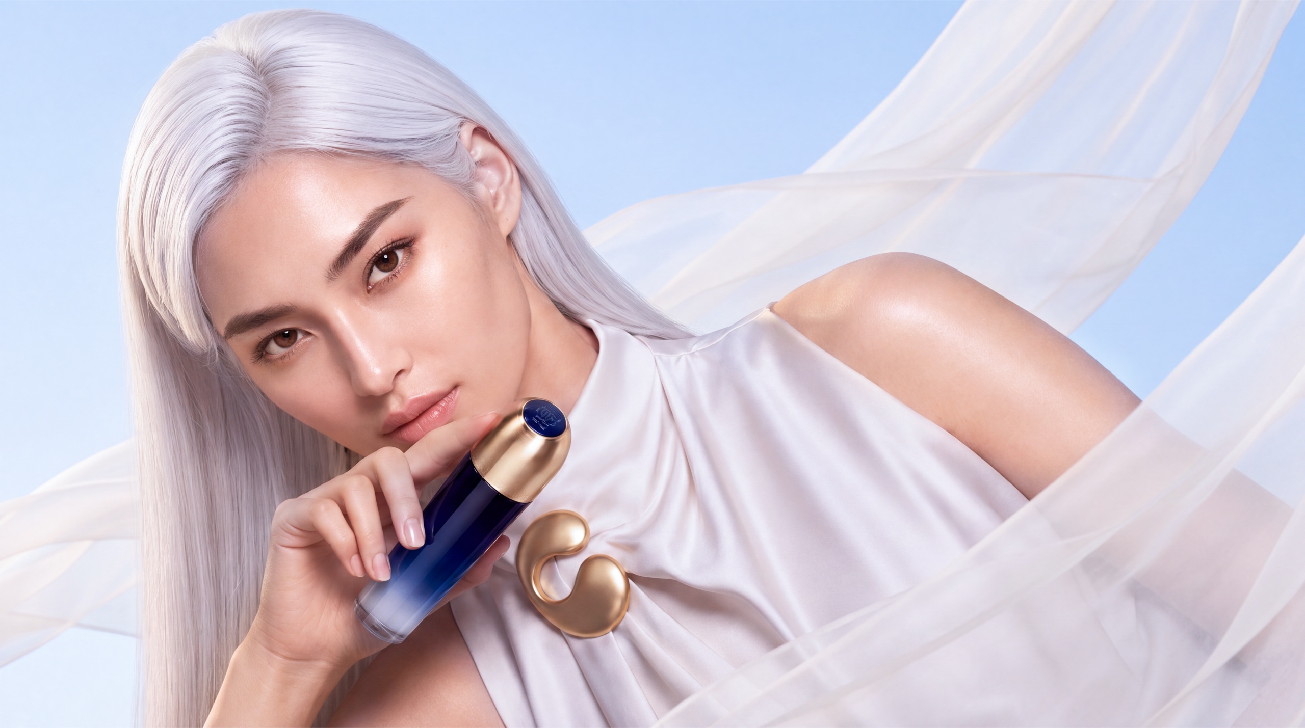 The muse holding the blue-and-gold beauty product among flowing ivory fabric